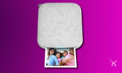 An HP Sprocket portable photo printer on a magenta background with a printed photo of a smiling family with two adults and two children.