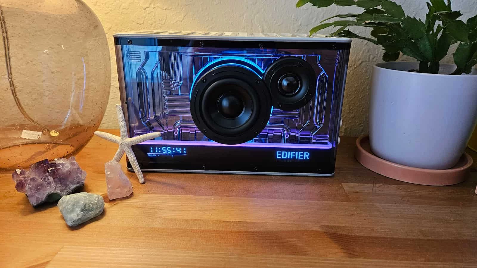A transparent Edifier QD35 Bluetooth speaker displaying its internal components is flanked by an amber glass vase, decorative stones, a starfish, and a potted plant.