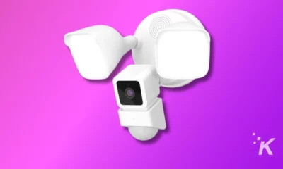 The image shows a modern white security camera system with multiple motion sensors on a vibrant purple background, representing smart home technology.