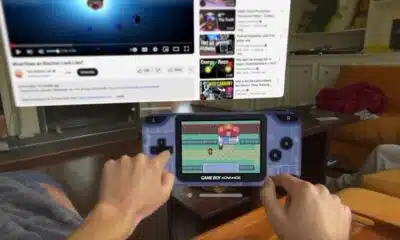 A person playing a game on a Game Boy Advance with a television displaying YouTube in the background; the couch and living room setting are slightly out of focus.
