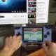 A person playing a game on a Game Boy Advance with a television displaying YouTube in the background; the couch and living room setting are slightly out of focus.