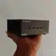 A person's hand holding a compact GEEKOM mini PC with visible USB ports, audio jack, HDMI port, and power button against a plain background.