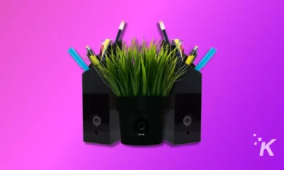 The image shows a black speaker with a pot of grass on top, surrounded by colorful pencils, against a gradient purple and pink background.