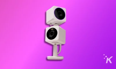 Two white security cameras stacked vertically against a vivid purple background. They have prominent lenses and appear modern. A subtle logo in the corner.