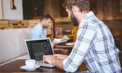 A man in a plaid shirt working on a laptop with analytical graphs on the screen, sitting at a wooden table in a cafe with a cup of coffee, with another person working in the background.