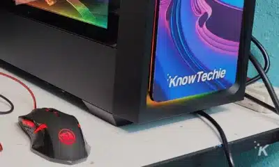 A gaming mouse with red accents connected to a computer with a monitor displaying the "KnowTechie" logo, on a white messy table against a teal wall.