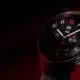 A smartwatch with a black interface displaying time, date, heart rate, and steps, set against a dark red background.