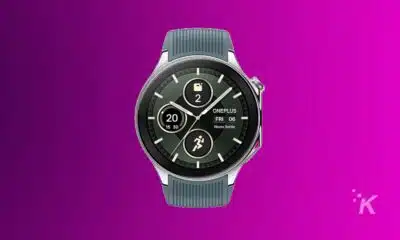 A OnePlus smartwatch with a black display featuring multiple dials showing the time, date, step count, and battery level against a purple background.