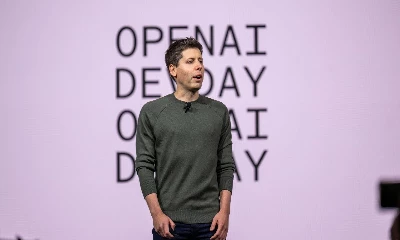 An individual is standing on stage with "OPENAI DEMO DAY" in the background, wearing a dark green sweater and dark pants, presenting to an audience.