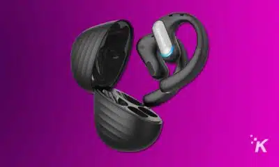Black wireless earphones with ergonomic hooks are emerging from a matching charging case against a vibrant purple background. They appear sleek and modern. OpenRock Pro headphones