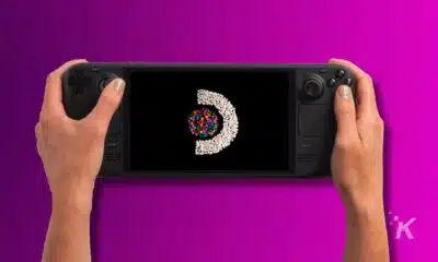 A pair of hands holding a steam deck oled gaming handheld with a colorful spiral galaxy on its screen, against a vibrant purple background.