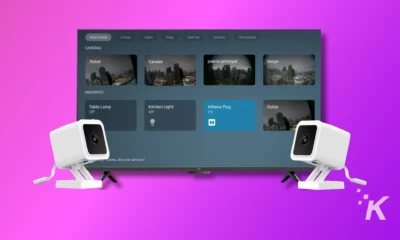 Two white security cameras are facing a colorful graphical user interface for a smart home system, which shows camera feeds and controls for various devices.