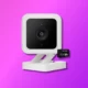 A Wyze security camera with a white frame, black lens, and a 32 GB microSD card against a vibrant purple background.